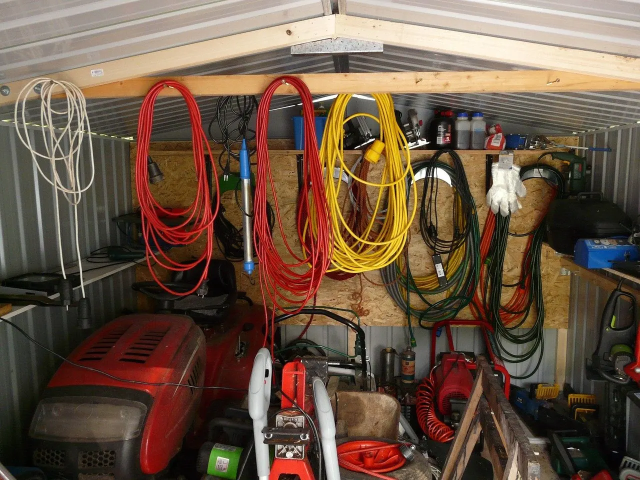 Gerätehaus aus Blech mit aufgehängten roten, gelben und grünen Verlängerungskabeln an der Wand, darunter Aufsitzmäher und Gartengeräte