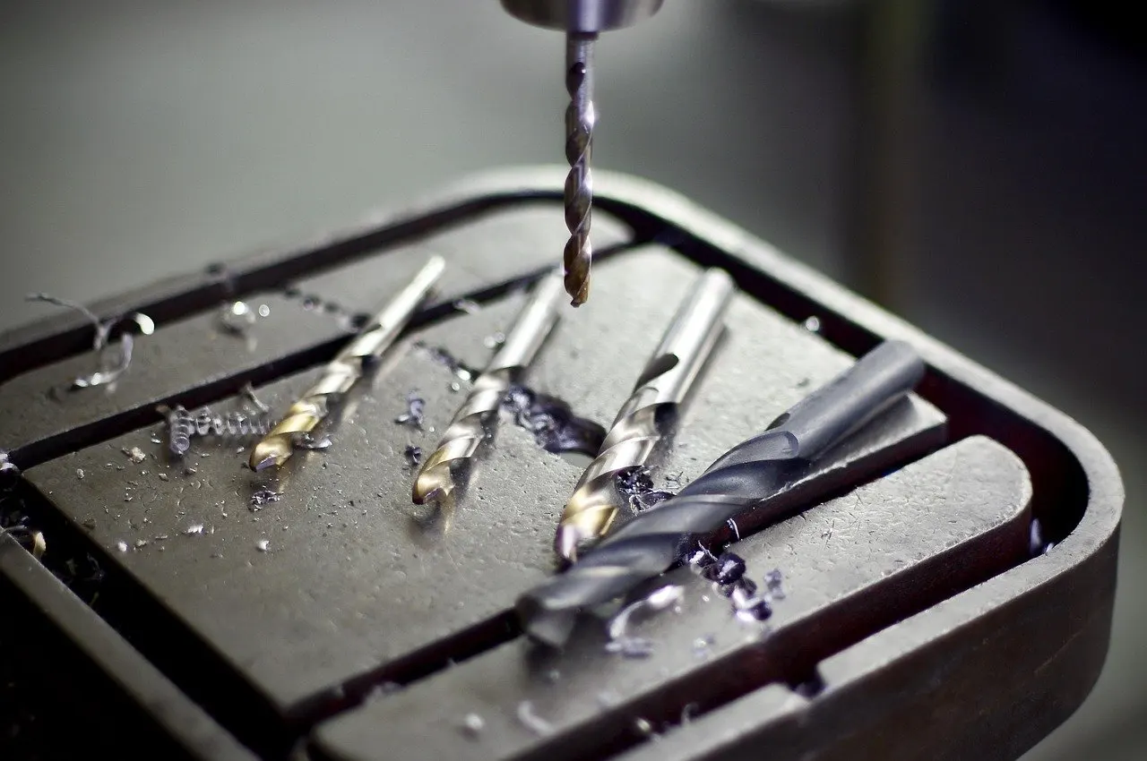 Tisch einer Ständerbohrmaschine mit vier Spiralbohrern und Metallspänen, darüber ein eingespannter Bohrer im Futter