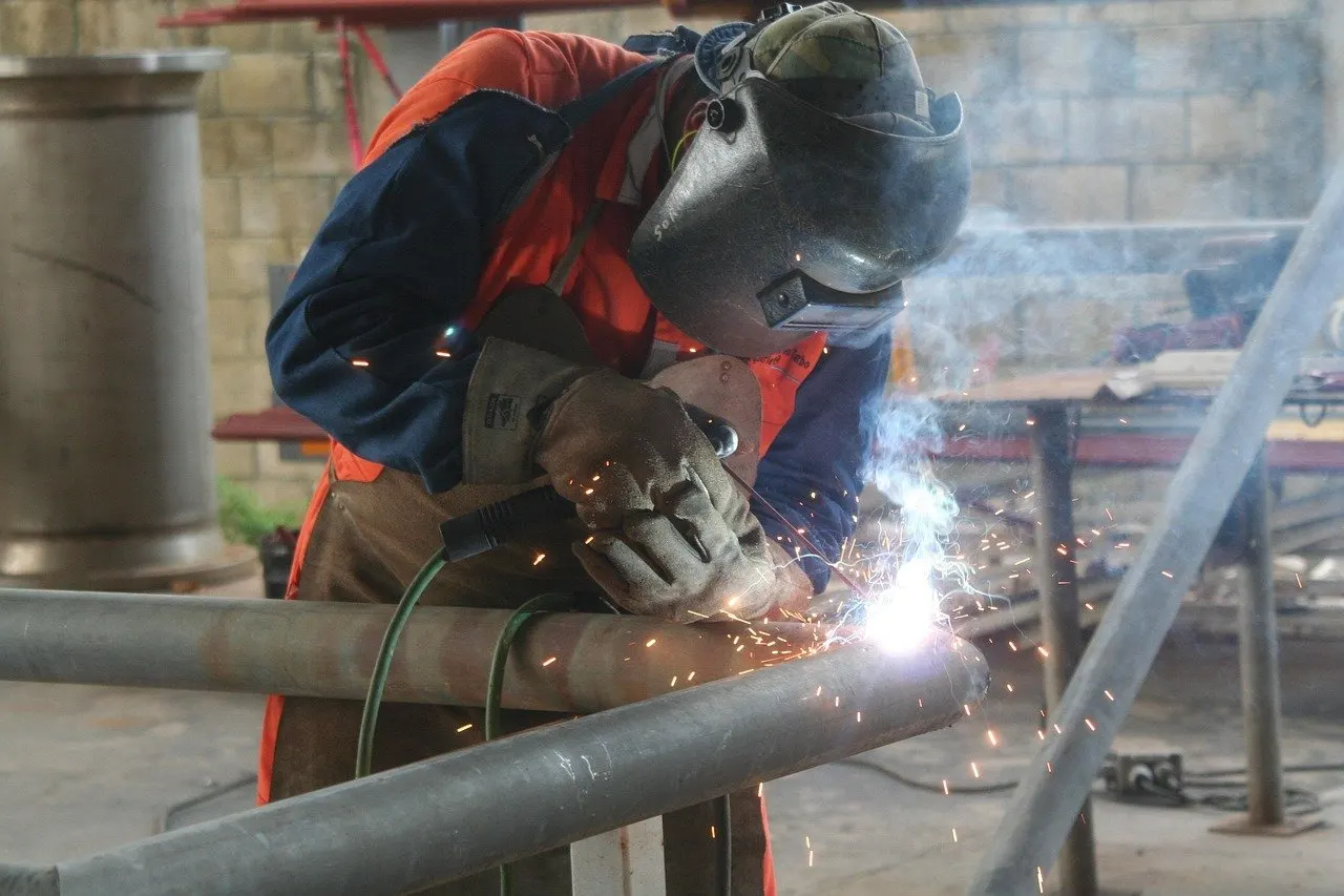 Schweißer mit Schweißhelm und Lederhandschuhen schweißt an einem Stahlrohr, Funken sprühen, eine Rauchfahne steigt auf