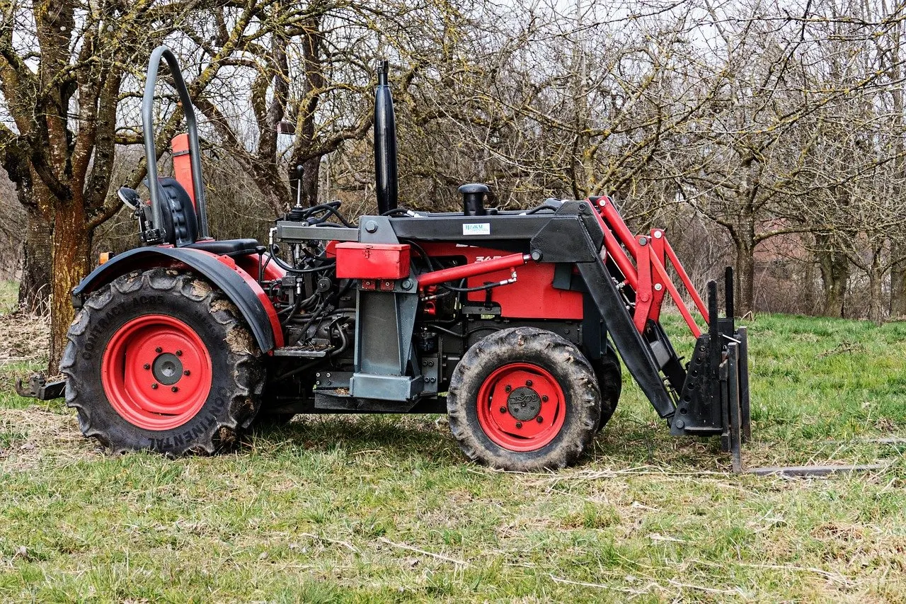 Roter Kleintraktor mit Überrollbügel und Frontlader samt Palettengabel steht auf einer Wiese zwischen kahlen Obstbäumen