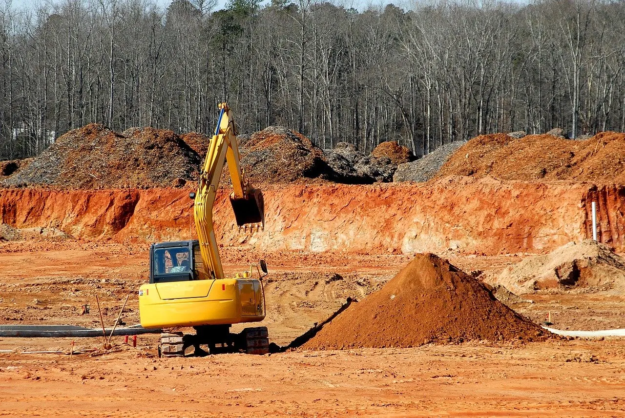 Gelber Kettenbagger mit angehobenem Löffel auf einer Erdbaustelle, daneben ein frisch aufgeschütteter Erdhaufen, dahinter eine Grubenkante und kahler Wald